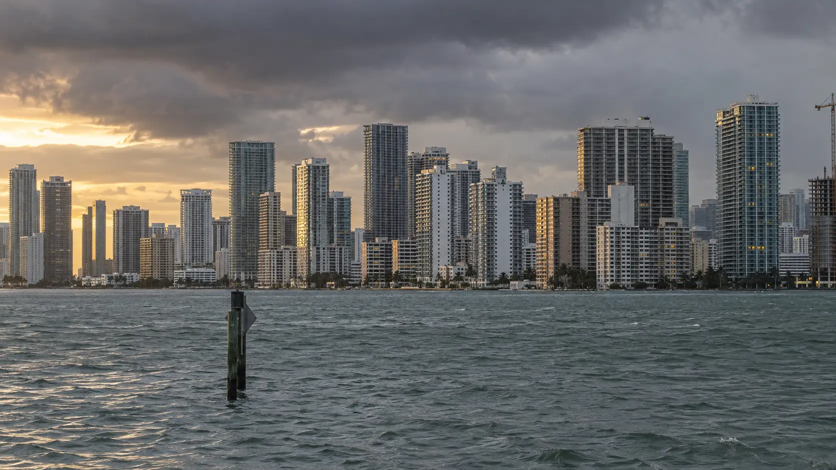 Condo towers along Biscayne Bay in Miami under rainy-season clouds, seen across choppy water