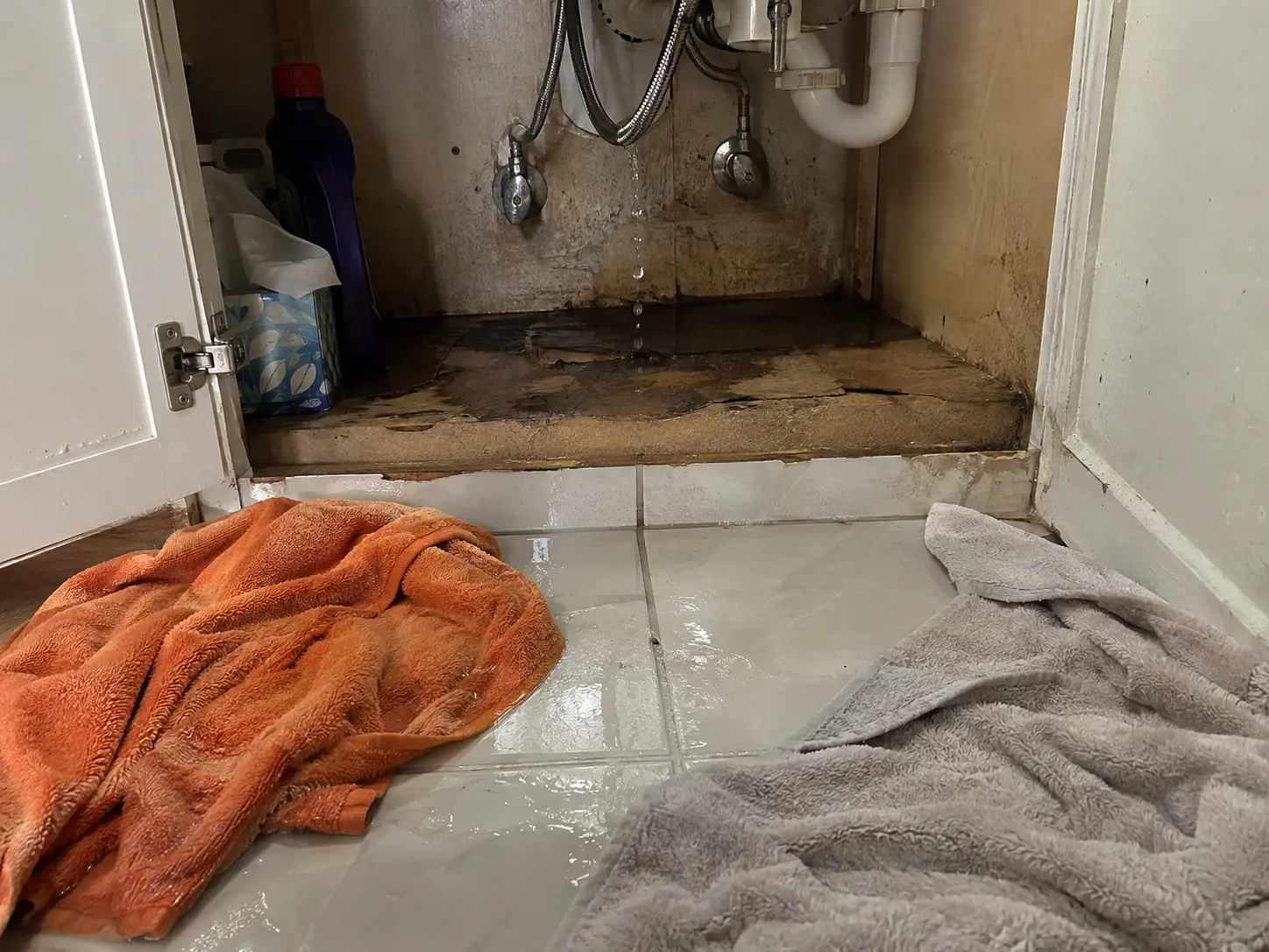 A burst supply line dripping inside a bathroom vanity onto a swollen cabinet floor, with towels on the wet tile in front